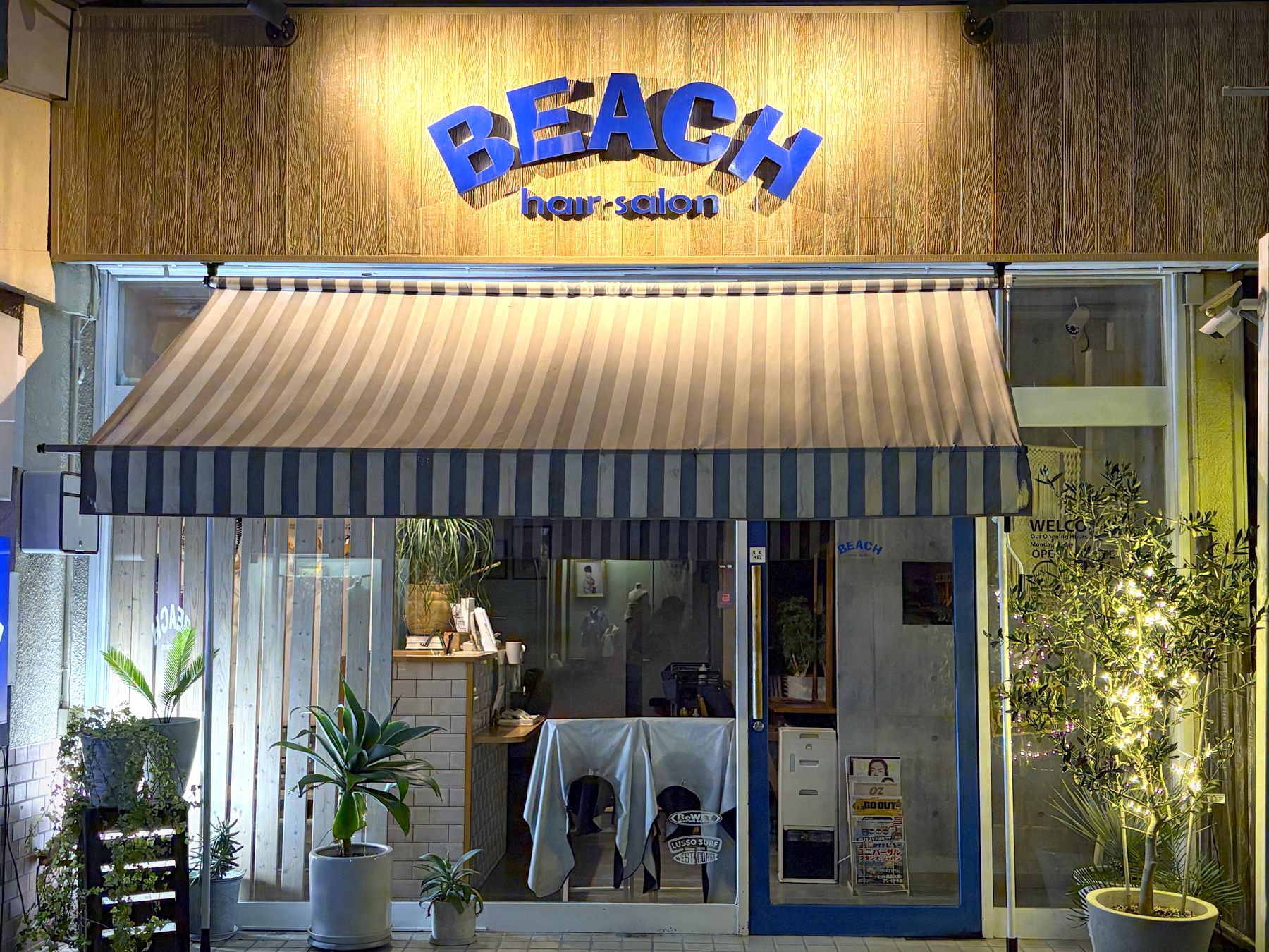 夜のhair salon BEACHの外観。青白ストライプのオーニングと、ブルーのBEACHサインに灯りがともる